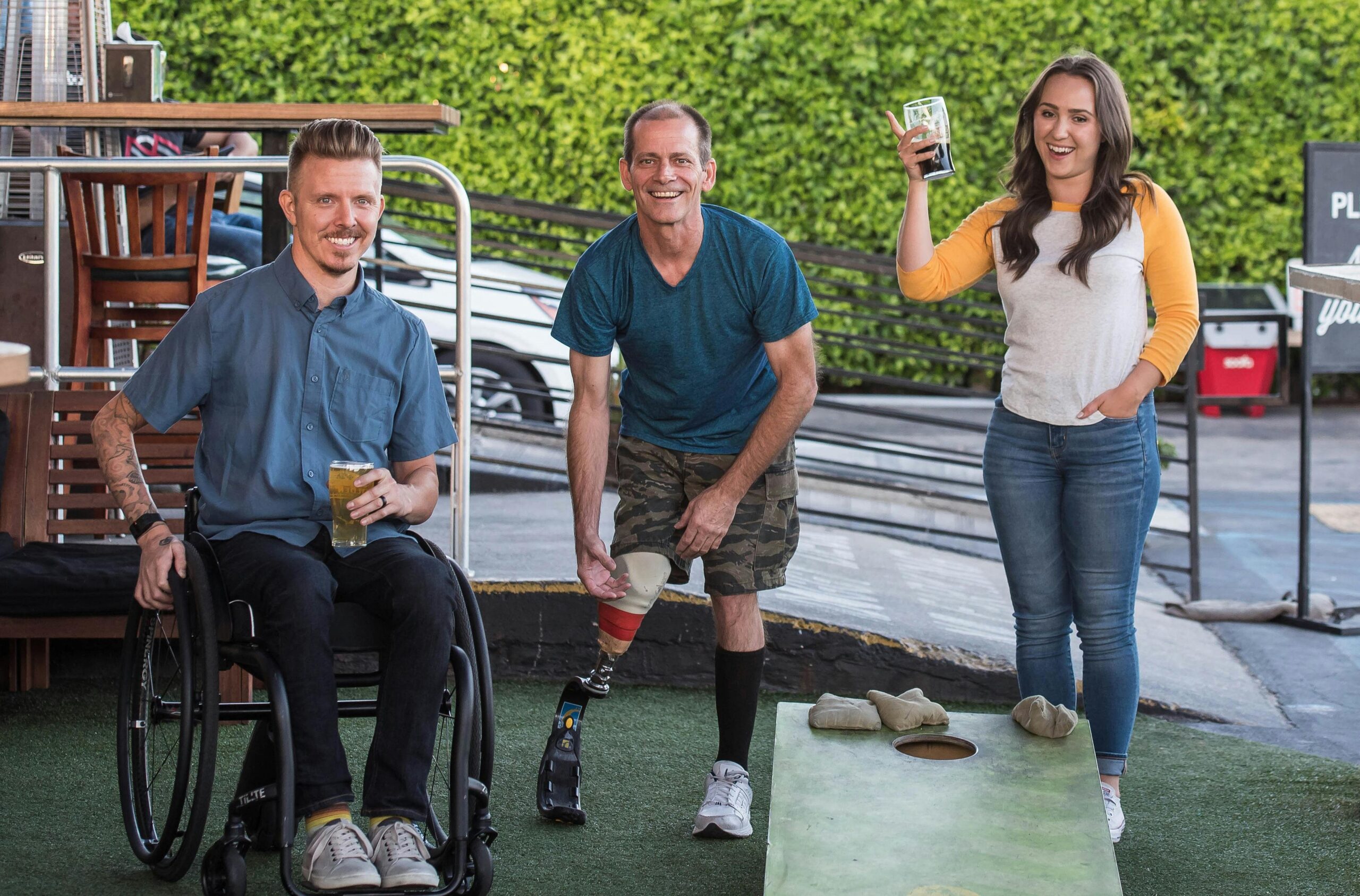 Three friends playing cornhole outdoors, enjoying drinks and smiling.